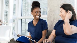 two female nurses in scrubs sitting on a couch and looking at a notebook while smiling and talking to each other